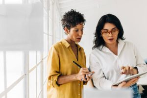 Two business women having a discussion, they're standing in an office and using a tablet. Professional women making a to do list as part of their project planning.