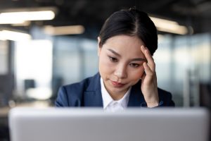 Asian businesswoman wearing a suit working on a laptop at the office, appearing stressed and worried. The image conveys concepts of business challenges, stress, and concentration